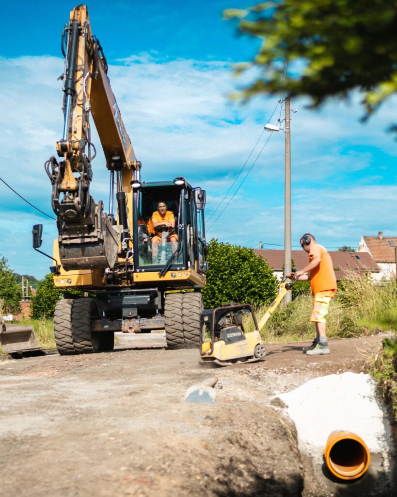 Baustellenszene auf einem unbefestigten Weg: Ein gelber CAT-Mobilbagger steht auf der Straße, während rechts ein Bauarbeiter in orangefarbener Arbeitskleidung und mit Gehörschutz eine Rüttelplatte bedient. Im Vordergrund ragt ein orangefarbenes Rohr aus einem Graben.