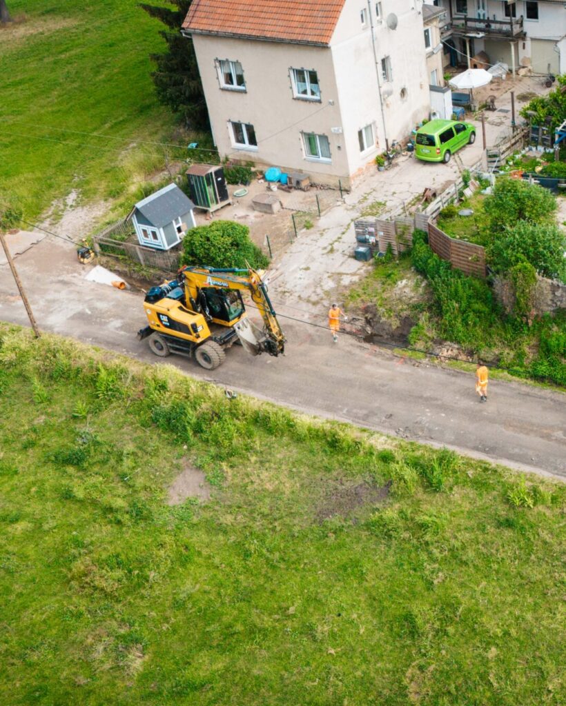Luftaufnahme einer Baustelle auf einer schmalen Dorfstraße. Zu sehen sind ein gelber Bagger, ein Transporter mit Pritsche, ein kleinerer Baustellenkipper, zwei Arbeiter in Warnkleidung sowie umliegende Wohnhäuser, Gärten und grüne Wiesen.