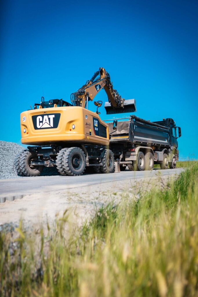 Gelber CAT-Mobilbagger belädt einen Lkw mit Schotter bei Straßenbauarbeiten im Freien