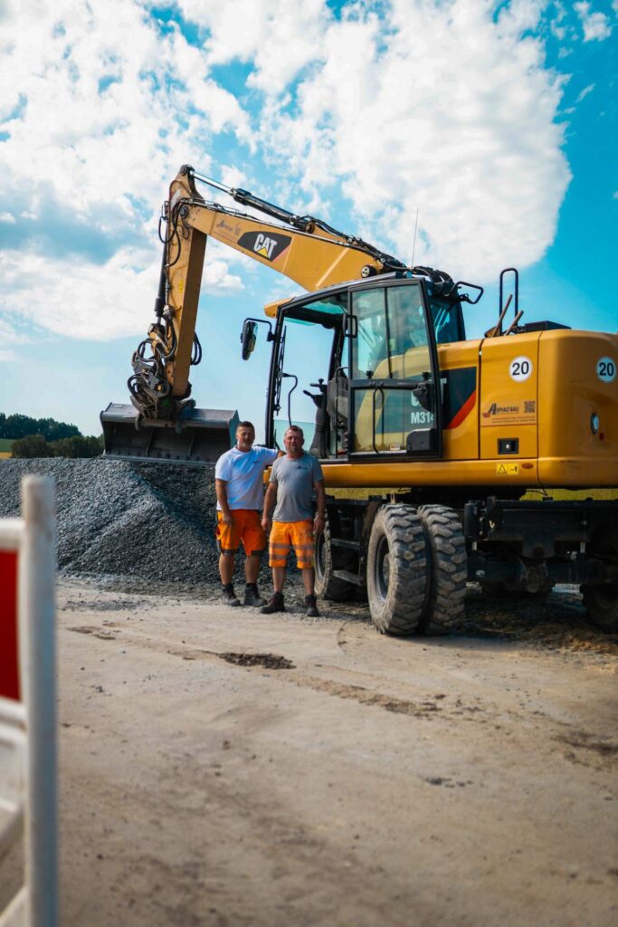 Zwei Bauarbeiter in Arbeitskleidung stehen lächelnd vor einem gelben CAT-Mobilbagger