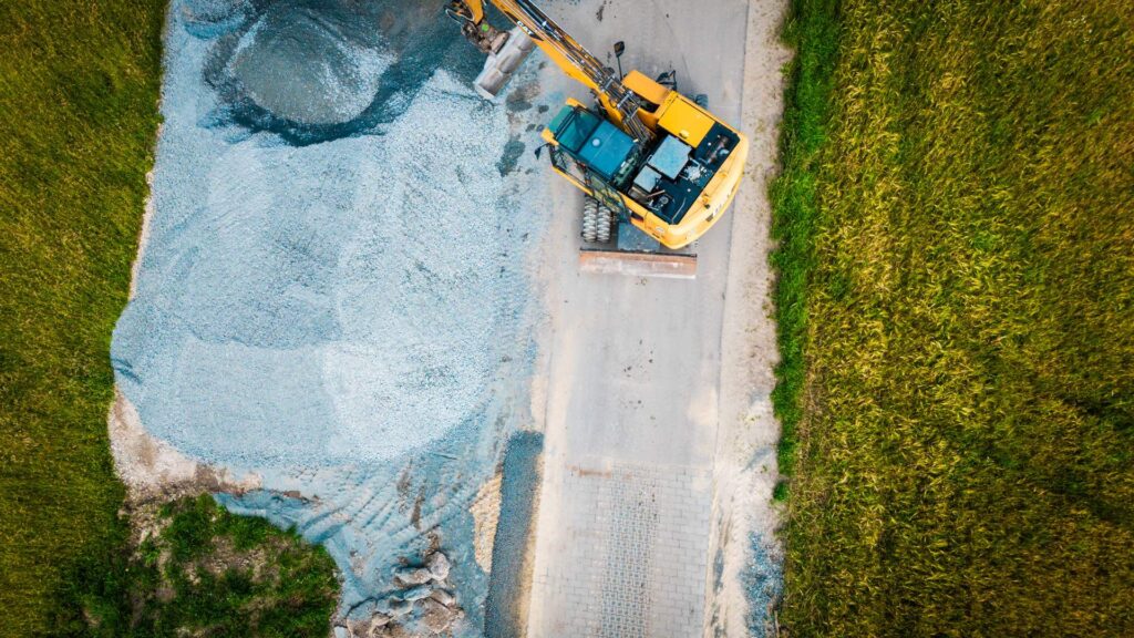 Vogelperspektive auf einen CAT-Bagger und ein großes Schotterdepot an einem Weg