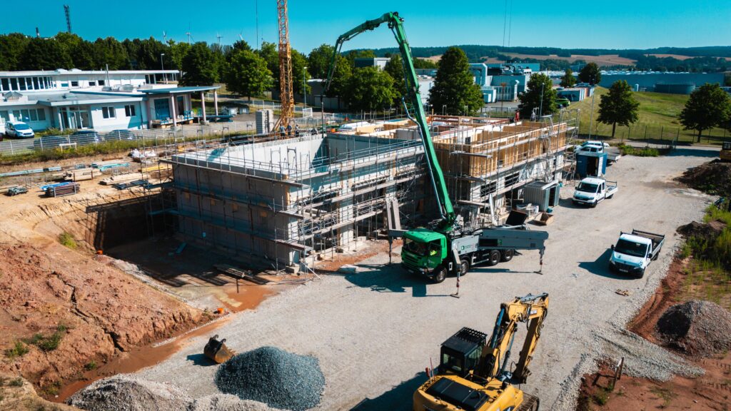 Luftaufnahme der Baustelle mit Betonpumpe, Bagger und Gewerbebau in Holz-Beton-Mischbauweise