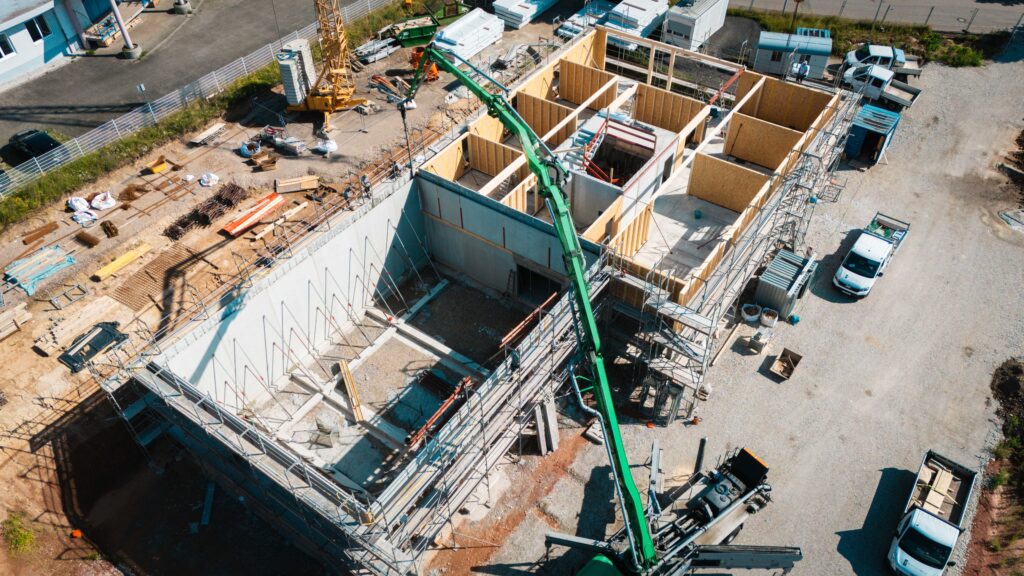 Drohnen-Schrägansicht auf den Rohbau mit Betonkeller, Holzwänden und Betonpumpe