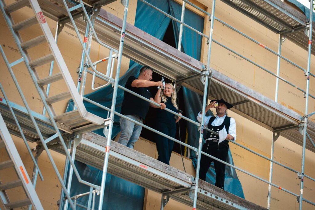 Zimmerer hält auf dem Baugerüst den Richtspruch mit erhobenem Glas neben der Bauherrschaft beim Richtfest.