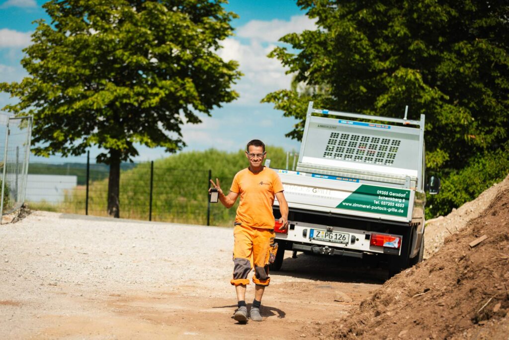 auarbeiter in oranger Arbeitskleidung macht das Peace-Zeichen vor dem Firmenwagen der Zimmerei Portack & Grille.