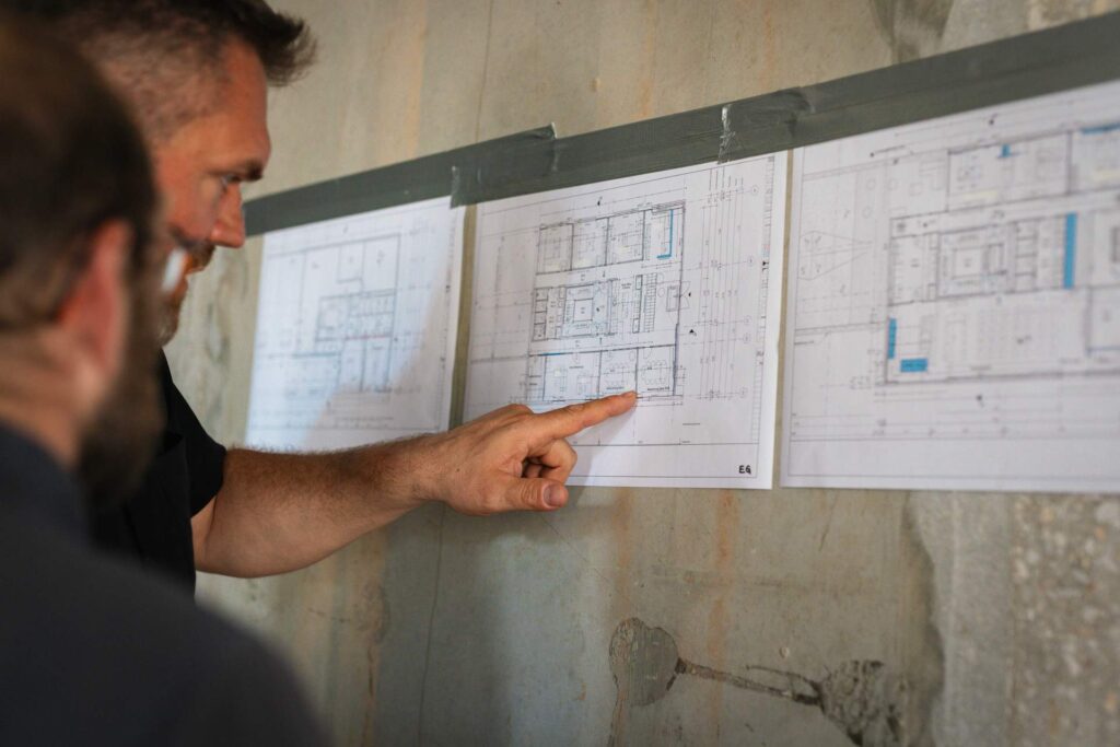 Mann zeigt im Rohbau auf den an der Wand befestigten Bauplan des neuen Gewerbegebäudes.