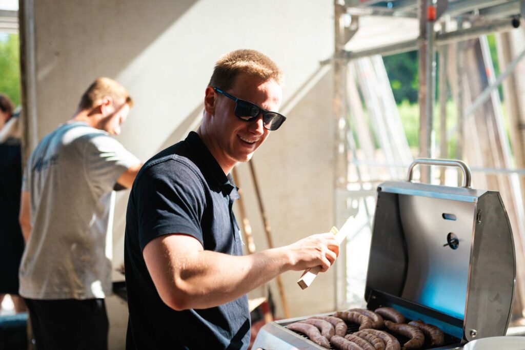 Mann mit Sonnenbrille steht lächelnd am Grill und bereitet Rostbratwürste für die Gäste des Richtfests zu.