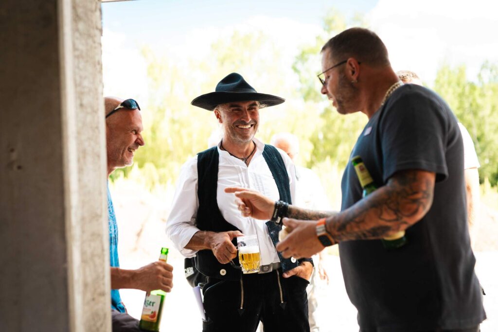 Zimmerer in Tracht unterhält sich fröhlich mit Bierglas in der Hand mit Gästen im Rohbau.