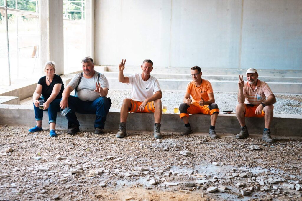 Fünf Mitarbeiter und Handwerker sitzen entspannt im Rohbau, trinken Bier und machen das Peace-Zeichen.