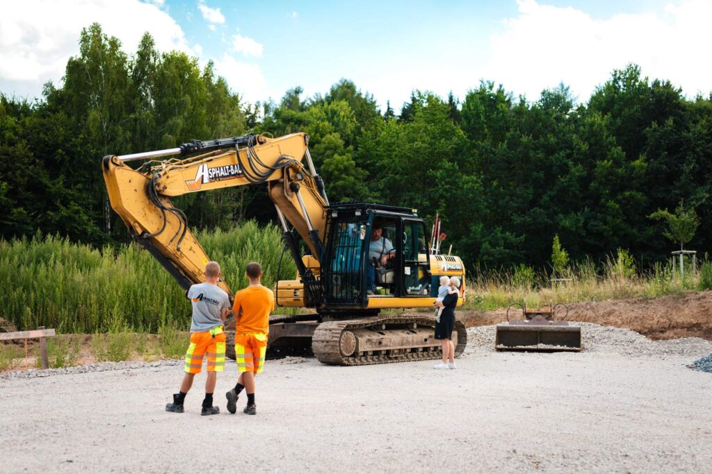 Gelber Bagger von Asphalt-Bau Chemnitz auf der Baustelle in St. Egidien mit Mitarbeitern im Vordergrund.
