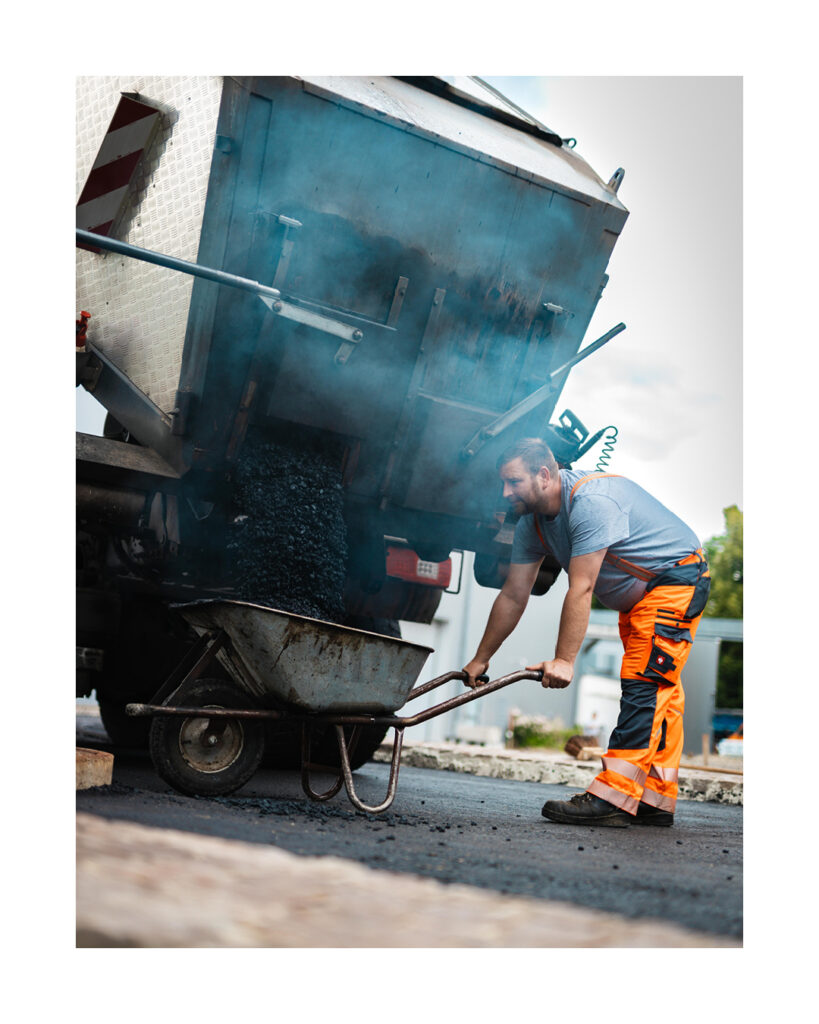 Seitliche Aufnahme eines Bauarbeiters, der eine Schubkarre unter die Ladeklappe eines LKW hält, während heißer, dampfender Asphalt herausfällt.
