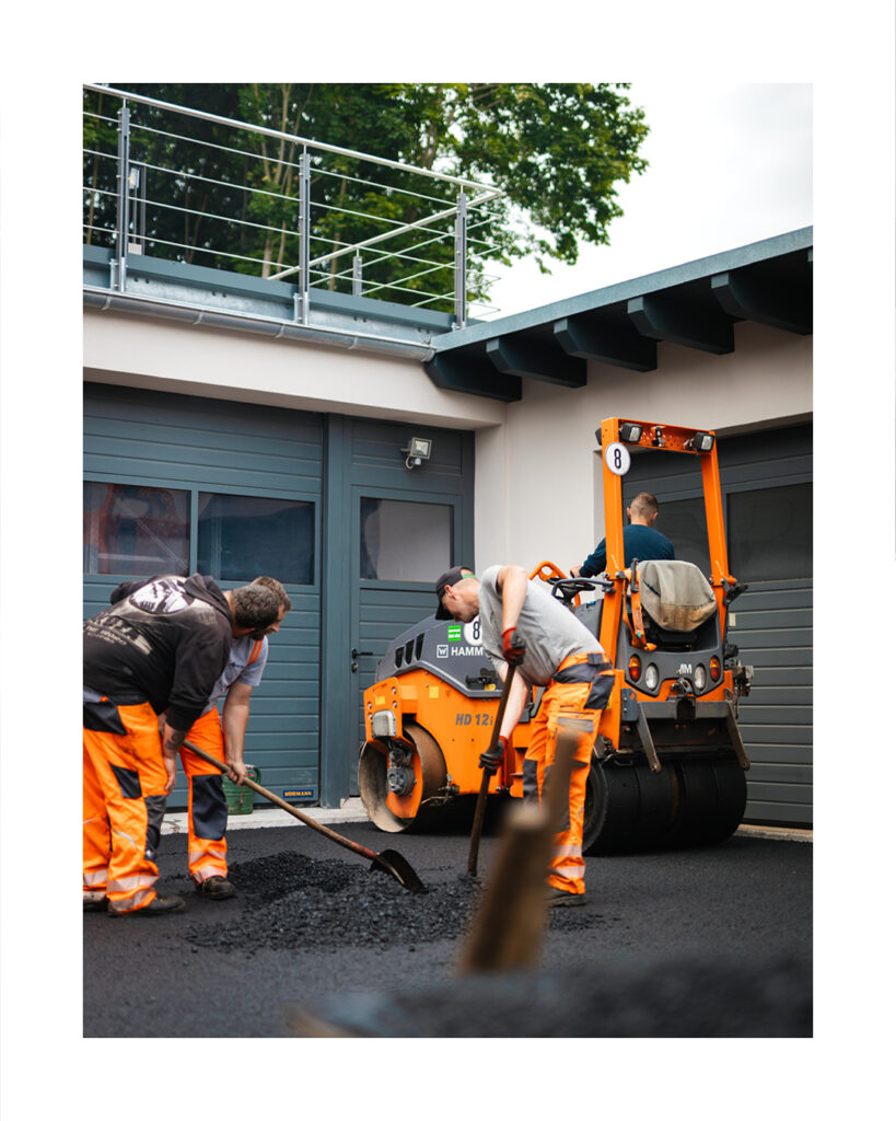 Baustellen-Foto von drei Straßenbauern in oranger Warnkleidung bei der Arbeit mit Schaufeln und einer kleinen Hamm HD 12 Walze. Die Szene spielt vor einer modernen Garagenanlage mit grauen Toren.