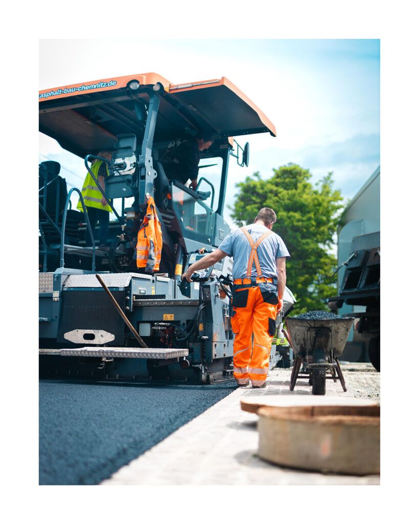 Detailaufnahme eines Bauarbeiters in oranger Latzhose, der von hinten den Vögele Straßenfertiger bedient. Rechts daneben eine Schubkarre mit Asphalt.