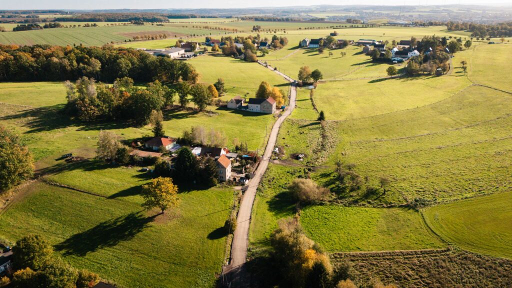 Luftaufnahme einer ländlichen Landschaft mit ausgedehnten grünen Wiesen, verstreuten Gehöften und einer Straße, die durch die Felder führt