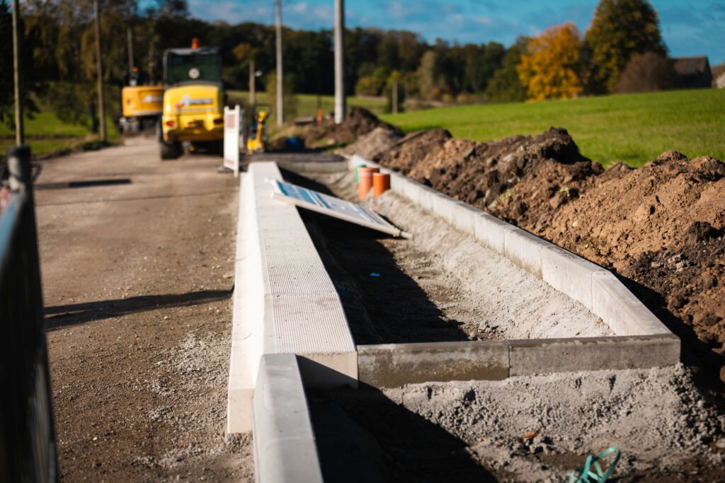 Baufstelle eines neuen Rad- oder Gehwegs mit Betonkanten und einem gelben Baufahrzeug im Hintergrund