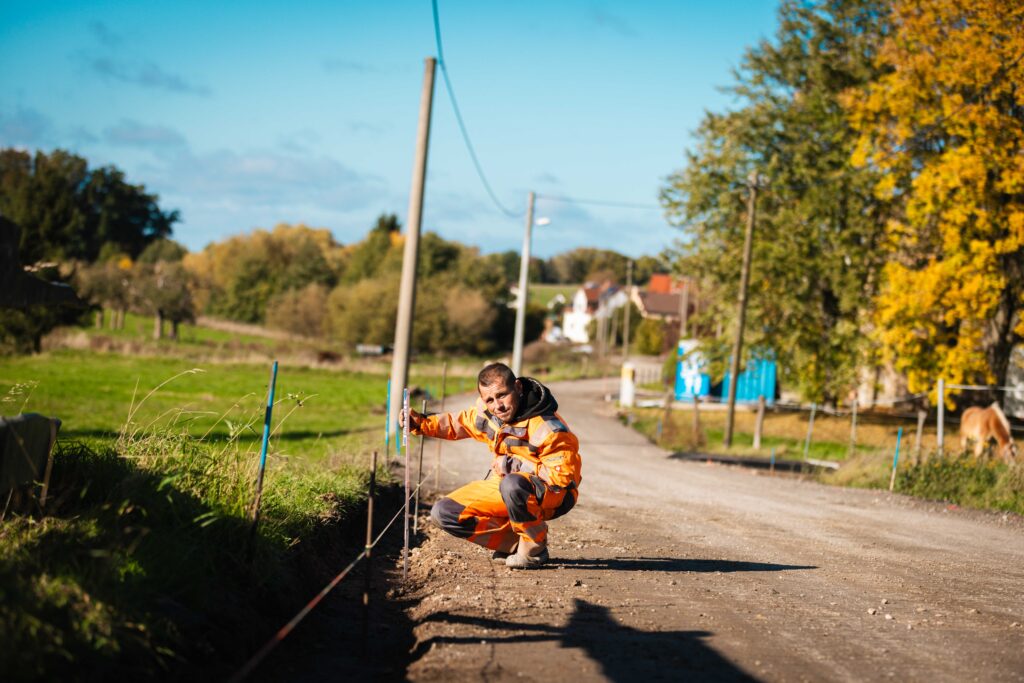 Ein Arbeiter in orangefarbener Schutzkleidung hockt am Rand einer unbefestigten Straße und hält eine Messlatte in den Boden