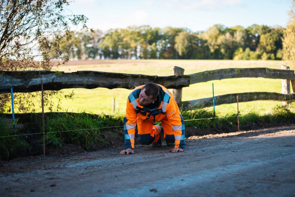 Ein Arbeiter in orangefarbener Schutzkleidung kniet auf einem Schotterweg und prüft den Boden neben einem Holzzaun
