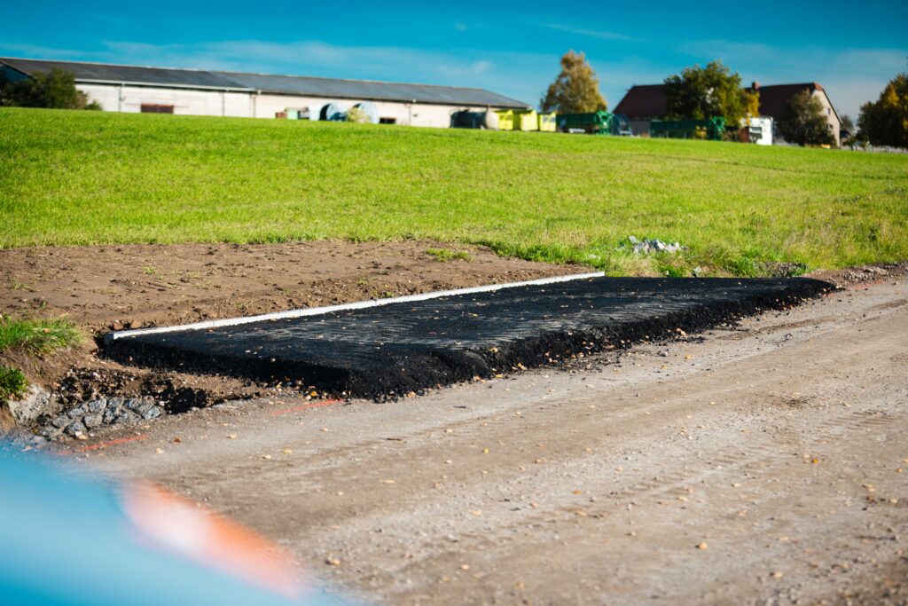 Frisch verlegter Asphaltbelag neben einer Erdstelle mit weißem Bordstein, im Hintergrund ein grünes Feld und landwirtschaftliche Gebäude