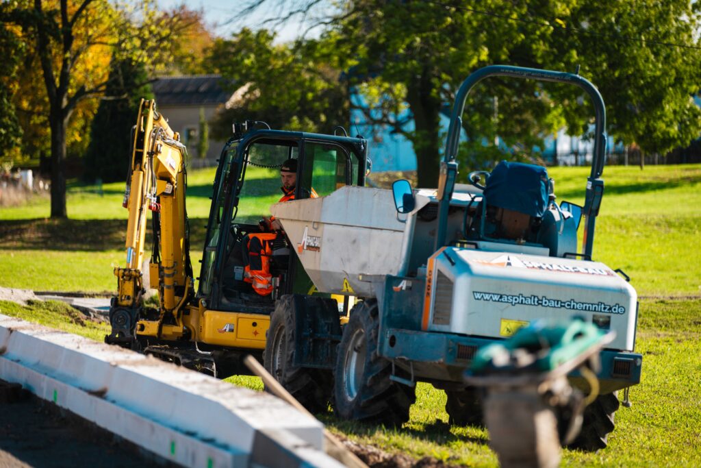 Ein Arbeiter bedient einen gelben Bagger. Daneben steht ein blau-weißes Fahrzeug mit 'Asphalt-Bau'. Im Vordergrund Betonkanten