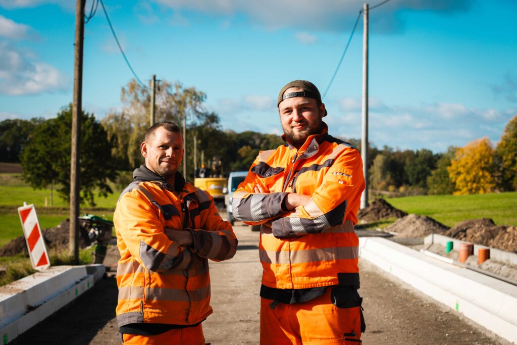 Zwei Männer in orangefarbener Schutzkleidung stehen mit verschränkten Armen auf einer Baustelle